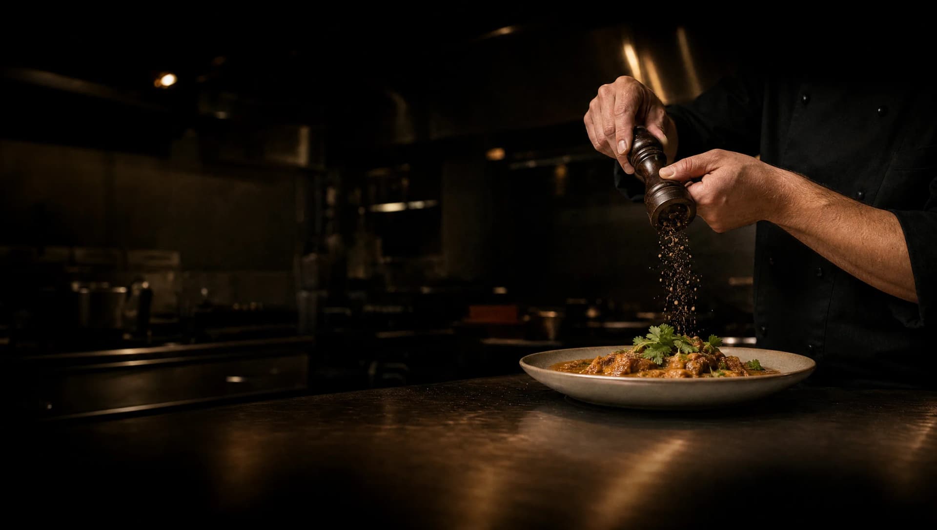 Chef sprinkling crushed black pepper over a plated dish on a stainless-steel kitchen pass